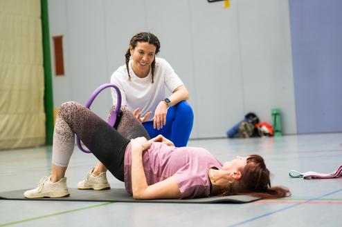 Zwei Frauen in Sportkleidung trainieren auf einer Matte, eine unterstützt die andere mit einem Fitnessgerät.