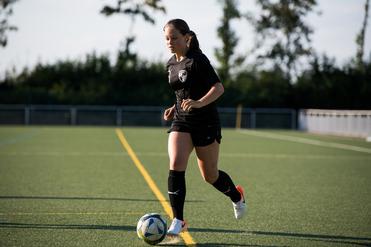 Eine Frau in schwarzer Sportkleidung dribbelt einen Fußball auf einem Kunstrasenplatz.