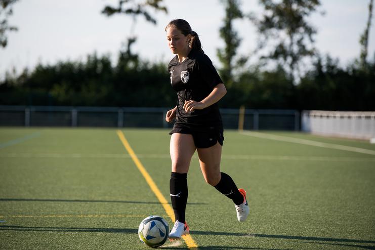 Eine Frau in schwarzer Sportkleidung dribbelt einen Fußball auf einem Kunstrasenplatz.