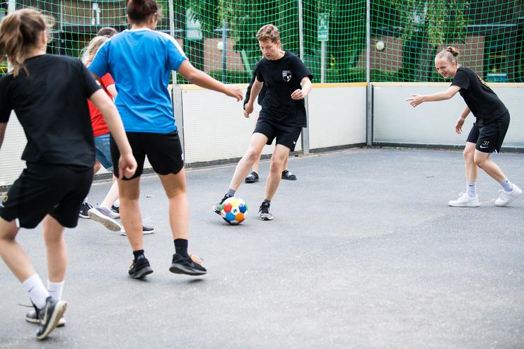 Gruppe von Jugendlichen spielt Fußball in einem umschlossenen Sportbereich mit Netz und Gummiuntergrund.