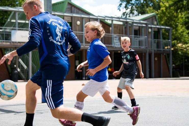Zwei Kinder in Sportkleidung und ein Trainer spielen Fußball auf einem Platz mit Gebäuden im Hintergrund.