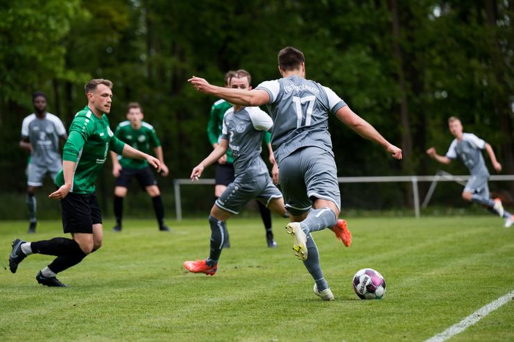Drei Fußballspieler in grünen und grauen Trikots auf einem Rasenplatz, während ein Spieler den Ball zuspielt.