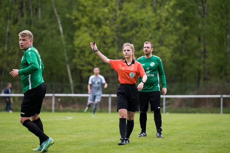 Schiedsrichterin in orangefarbenem Trikot zeigt eine Entscheidung, während zwei Spieler auf dem Fußballfeld stehen.