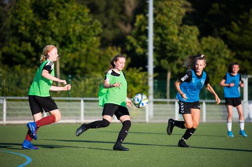 Drei Fußballspielerinnen in Trainingswesten schießen einen Ball auf einem Kunstrasenplatz unter blauem Himmel.