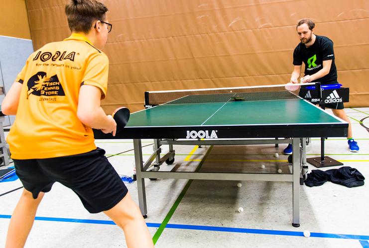 Zwei Tischtennisspieler trainieren an einem Tischtennistisch in einer Sporthalle, mehrere Bälle liegen auf dem Boden.