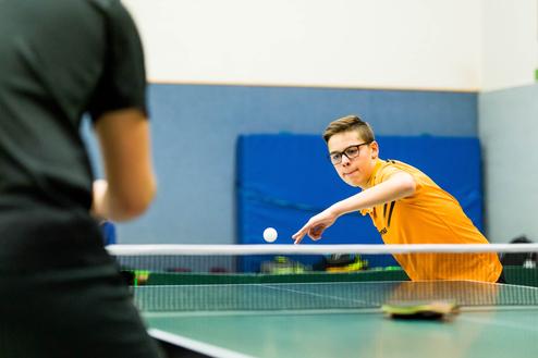 Junger Spieler in gelbem Trikot schlägt einen Tischtennisball über das Netz auf einer Tischtennisplatte.
