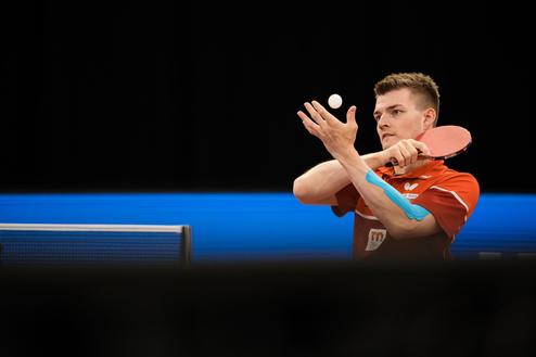 Spieler in rotem Shirt mit Tischtennisschläger hebt einen Tischtennisball vor einem blauen Hintergrund.