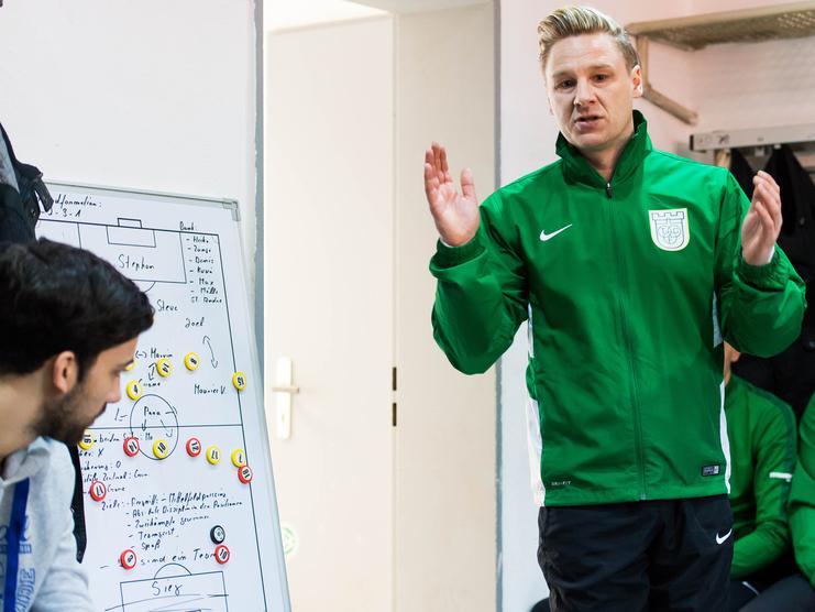 Trainer erklärt Taktik an Taktiktafel, Spieler hören im Hintergrund aufmerksam zu, in Umkleidekabine mit grüner Kleidung.